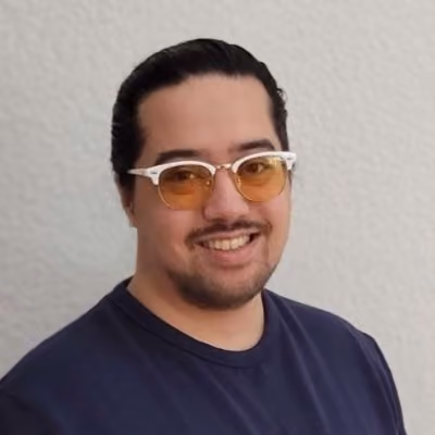 A smiling young man with short dark hair and a beard, wearing orange-tinted clear-framed clubmaster-style sunglasses and a navy blue t-shirt, photographed against a light textured wall.