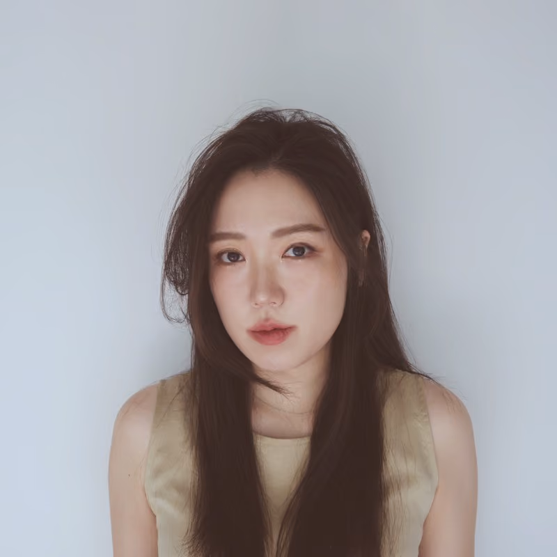 A young woman with long straight dark brown hair, wearing a sleeveless light olive/gold top, looking directly at the camera with a calm expression against a soft light gray backdrop.