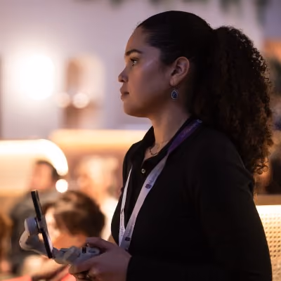 A young woman with a curly ponytail, wearing a black blazer and a lanyard with a conference badge, holds a tablet or device while attentively looking to the side at an event or conference.