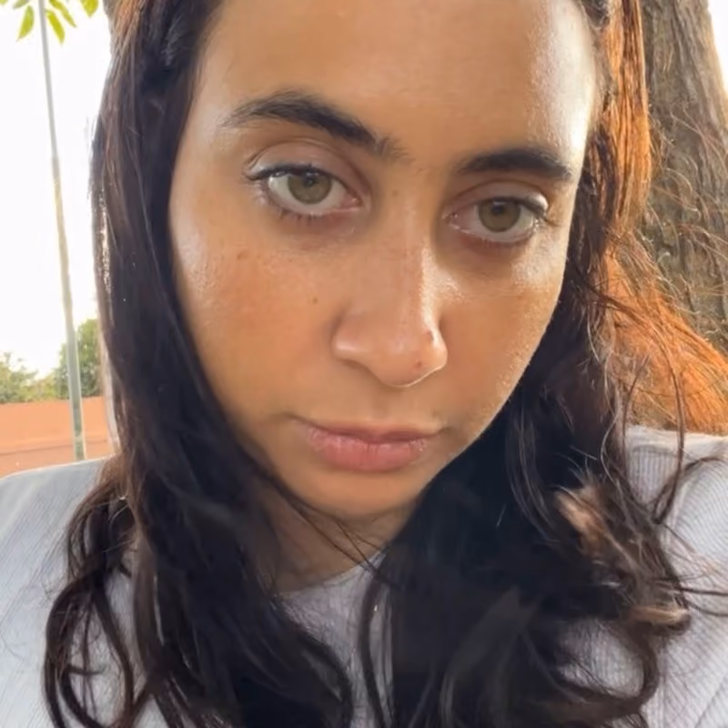 Close-up photo of a young woman with long dark wavy hair, green eyes, and a neutral expression, wearing a light gray top, with trees visible in the blurred outdoor background.