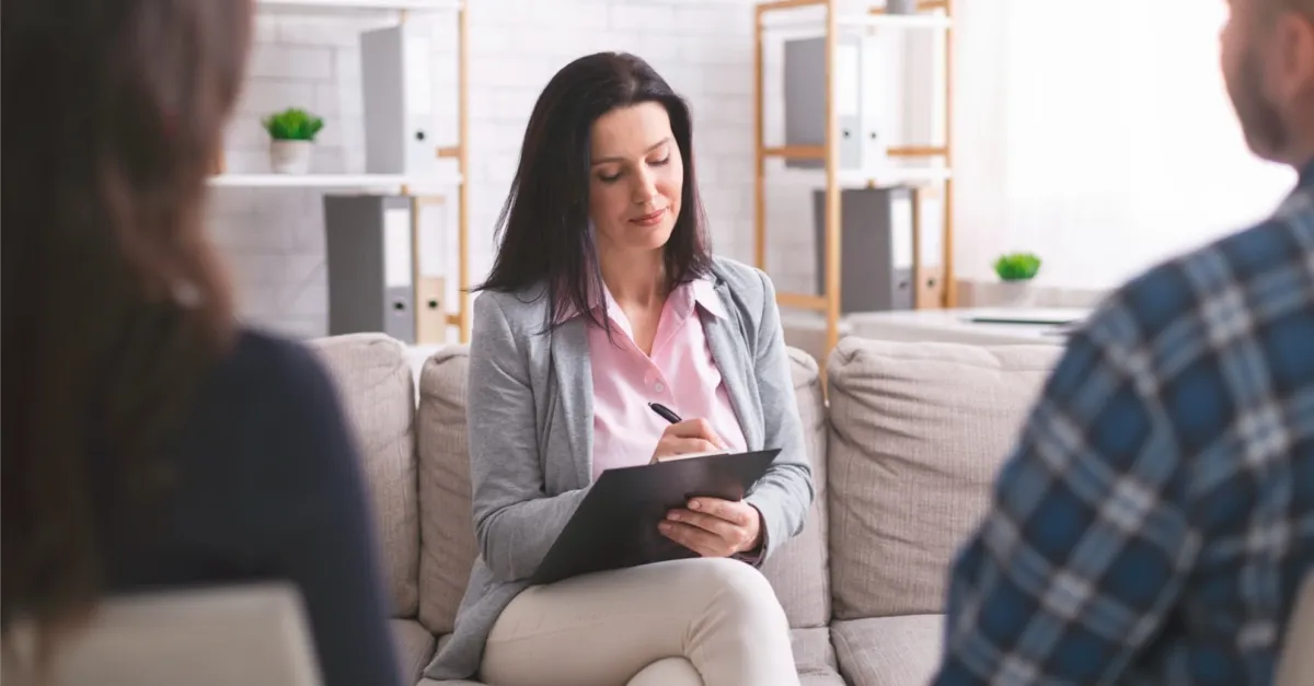 A woman therapist with a clipboard listens to two people during a counseling session.