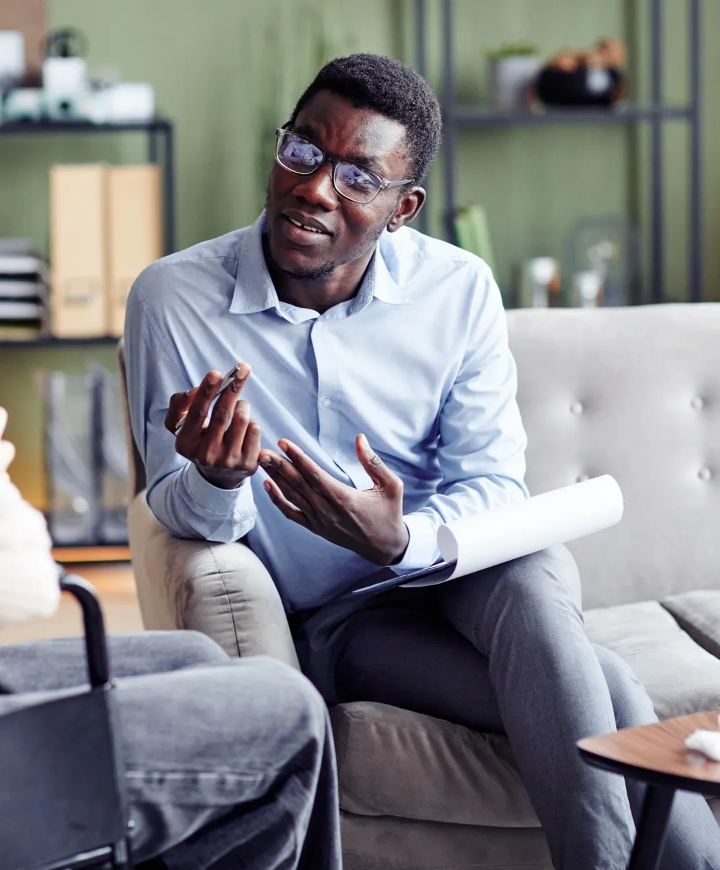Man in glasses and blue shirt talking and gesturing while sitting on a couch, holding papers.