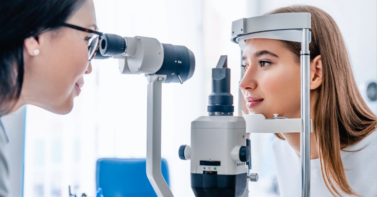 Optometrist examining a woman's eyes using a slit lamp in a bright, clinical setting.