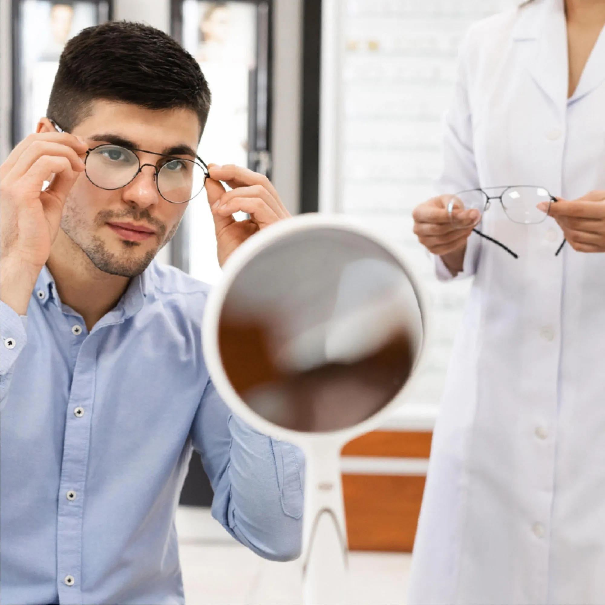 Man trying on round eyeglasses in front of a mirror with optometrist holding another pair nearby.