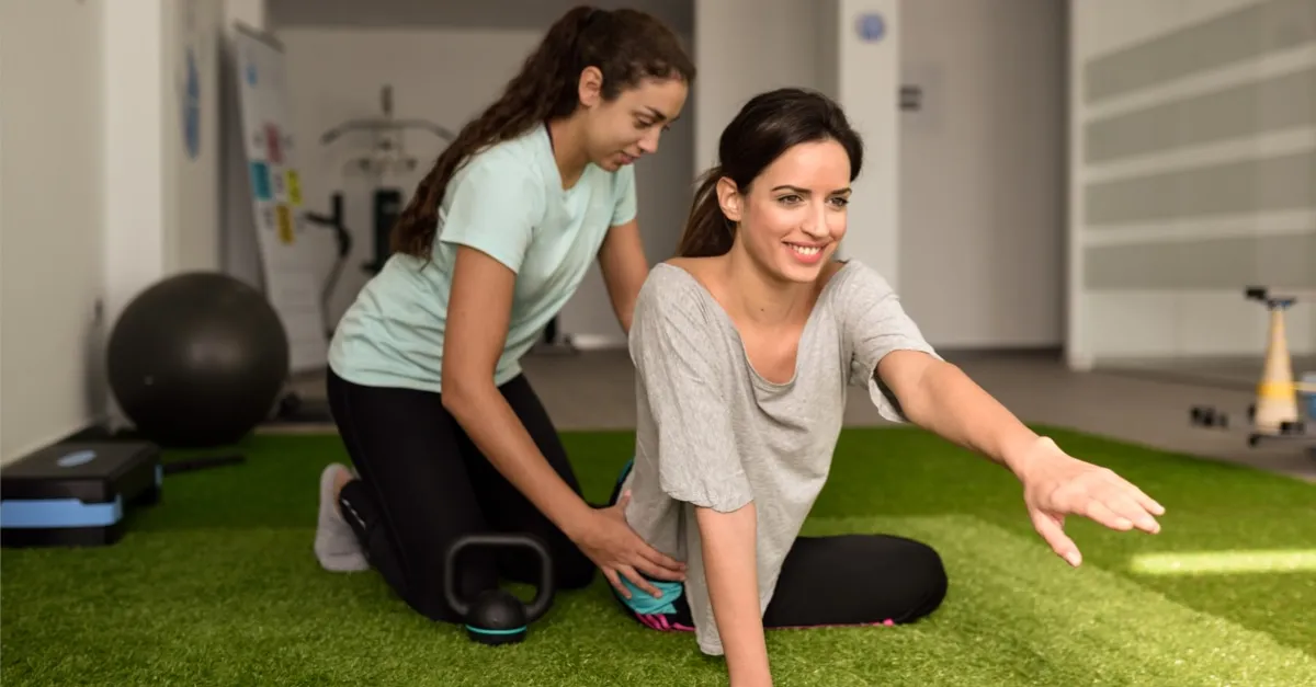 Physical therapist assists woman in stretching exercise on a green mat in a therapy room.