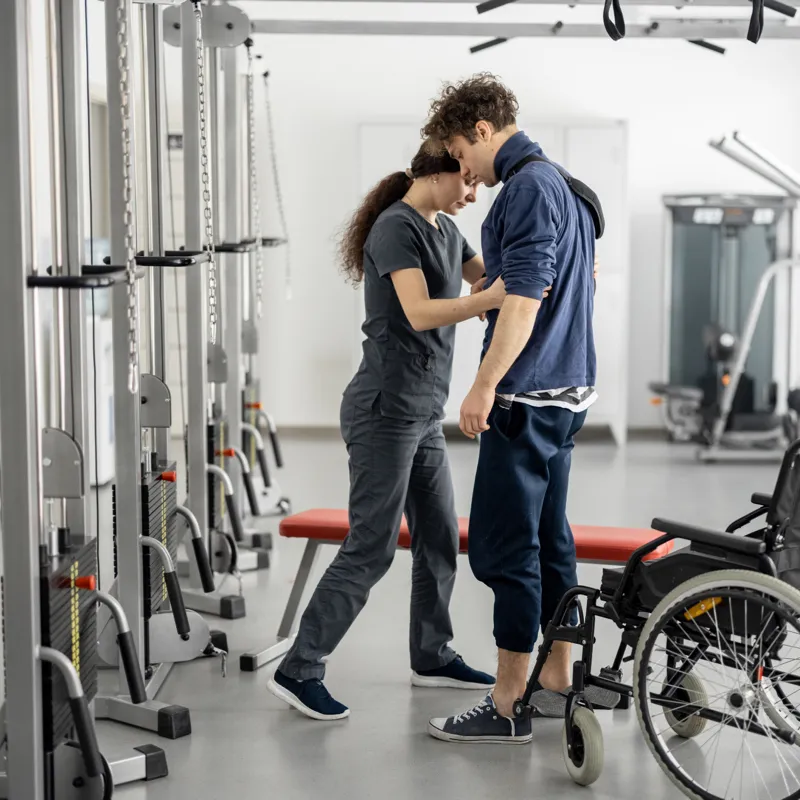 Physical therapist assists man standing up from wheelchair in a gym with exercise equipment.