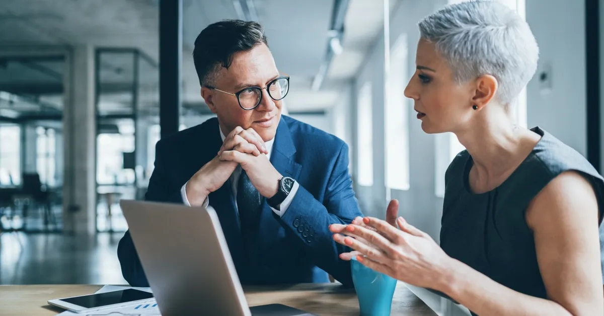 Two professionals in an office discussing work with a laptop and tablet on the table.