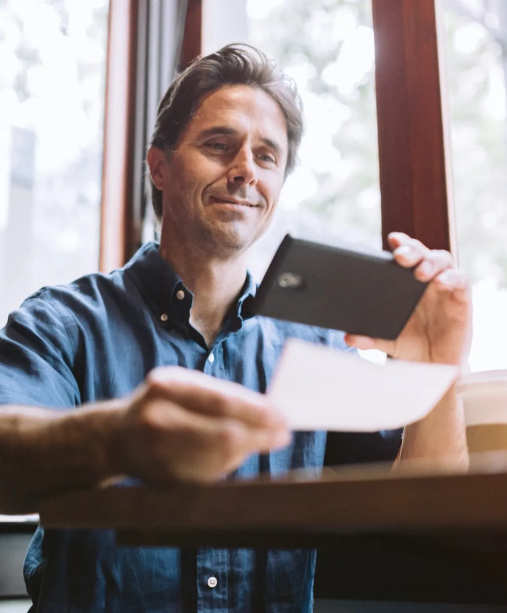 Man in a blue shirt sitting by window, smiling while using smartphone to scan a paper document.