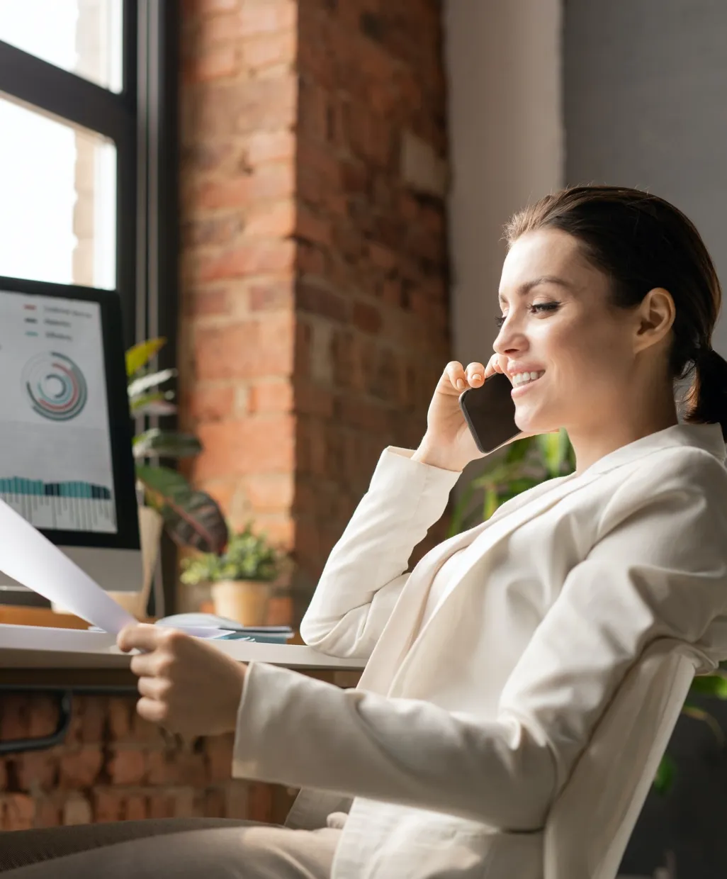 Woman in a white blazer talks on phone while reviewing papers at her desk with computer and plants nearby.