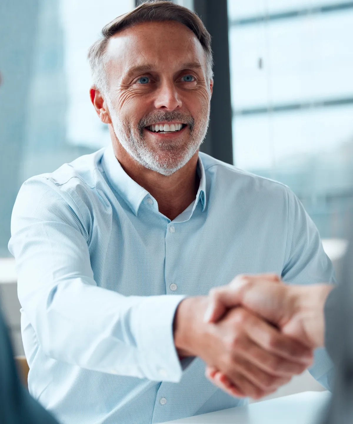Smiling middle-aged man in a light blue shirt shaking hands with someone across a table.