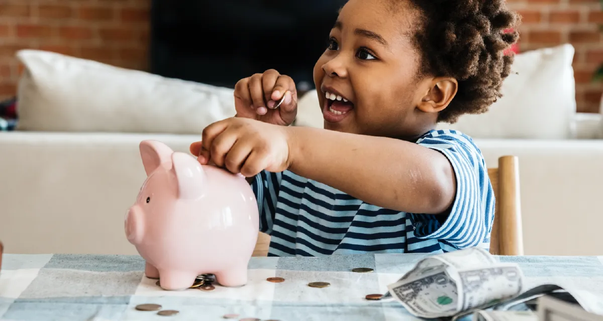 Smiling child putting a coin into a pink piggy bank on a table with scattered coins and dollar bills.