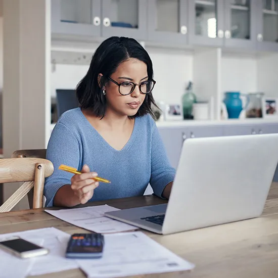 Woman in glasses and blue sweater working on laptop with documents and calculator on table.