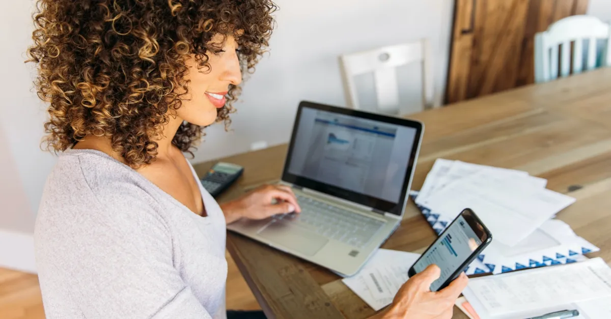Woman with curly hair using a laptop and checking financial data on her smartphone at a wooden table.