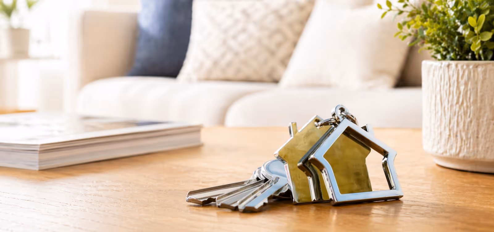 Silver house-shaped keychains with keys on a wooden table in a bright living room.