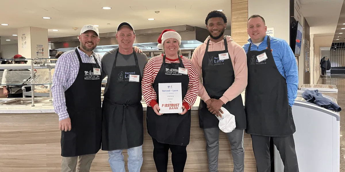 Five people wearing aprons smile indoors, one holding a sign saying Served with Love dinner by Firstrust Bank.