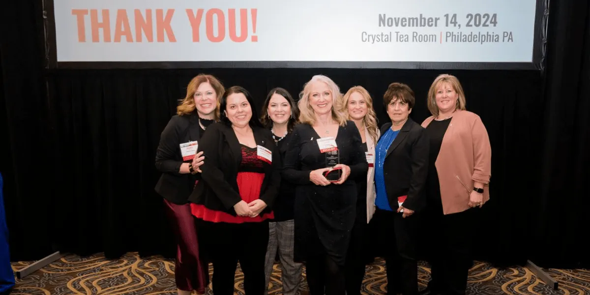 Seven smiling women at an award event with a 'Thank You' banner and event details in background.