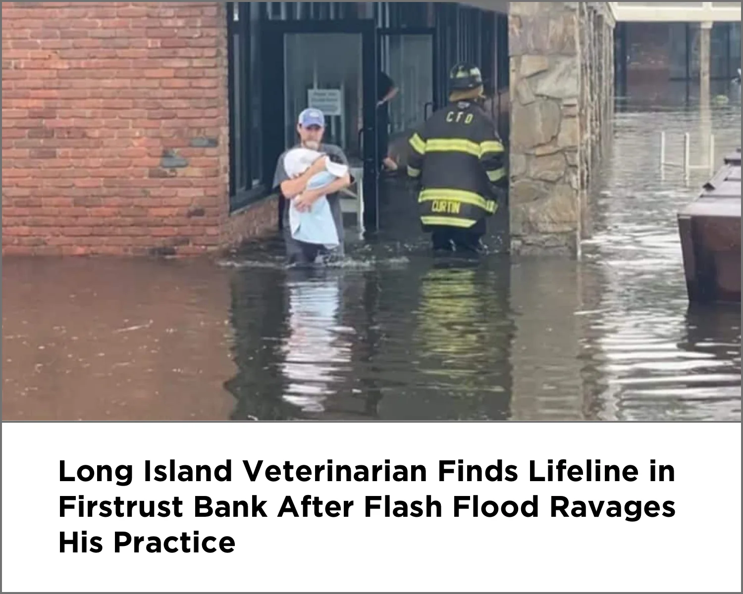 Man carrying a wrapped baby walking through floodwater outside a building with a firefighter nearby.