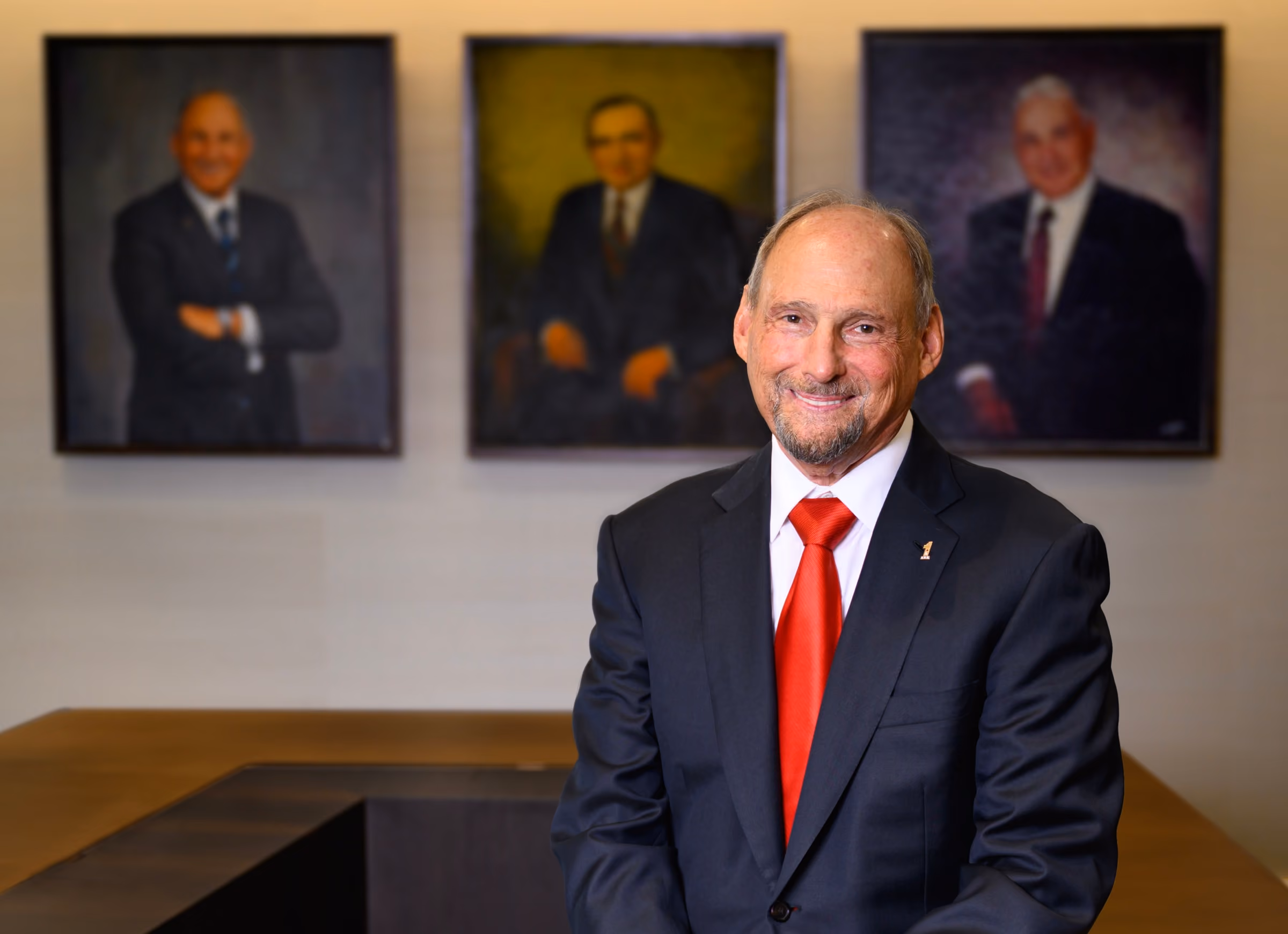 Smiling man in black suit and red tie standing in front of a table with three framed portraits behind him.
