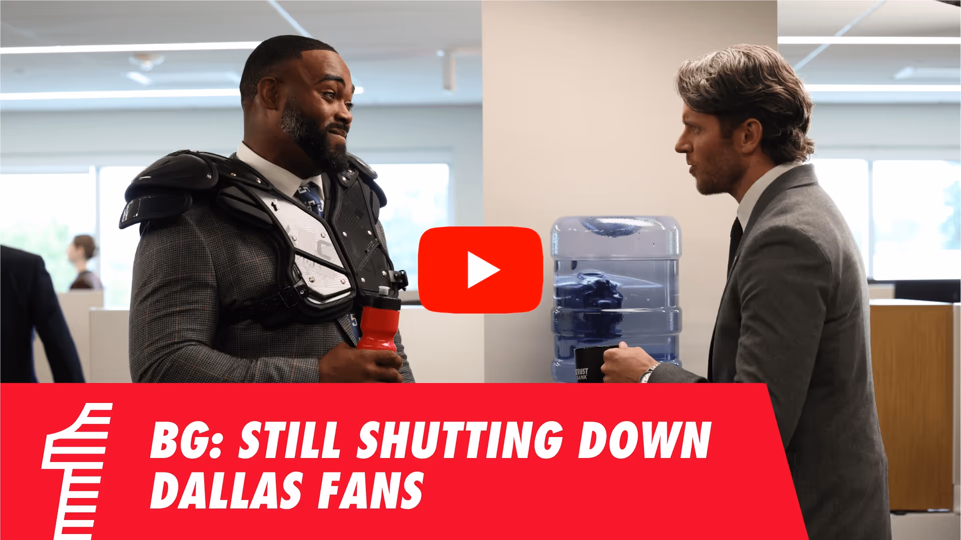Two men in office talking by water cooler, one wearing football shoulder pads and suit.