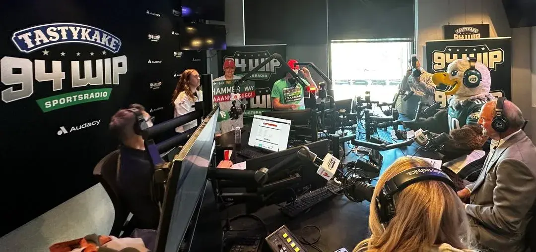Radio studio with hosts and a mascot wearing large headphones during a live broadcast.
