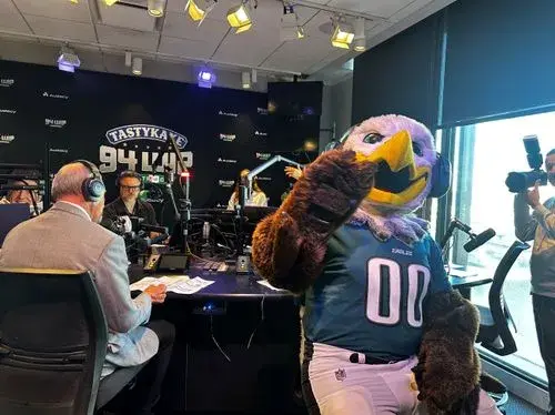 Eagles mascot giving thumbs up inside radio station studio during broadcast with hosts and cameraman.