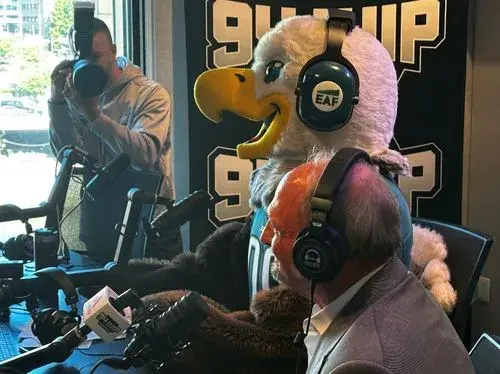 Man in a suit and a person in an eagle mascot costume wearing headphones in a radio studio.