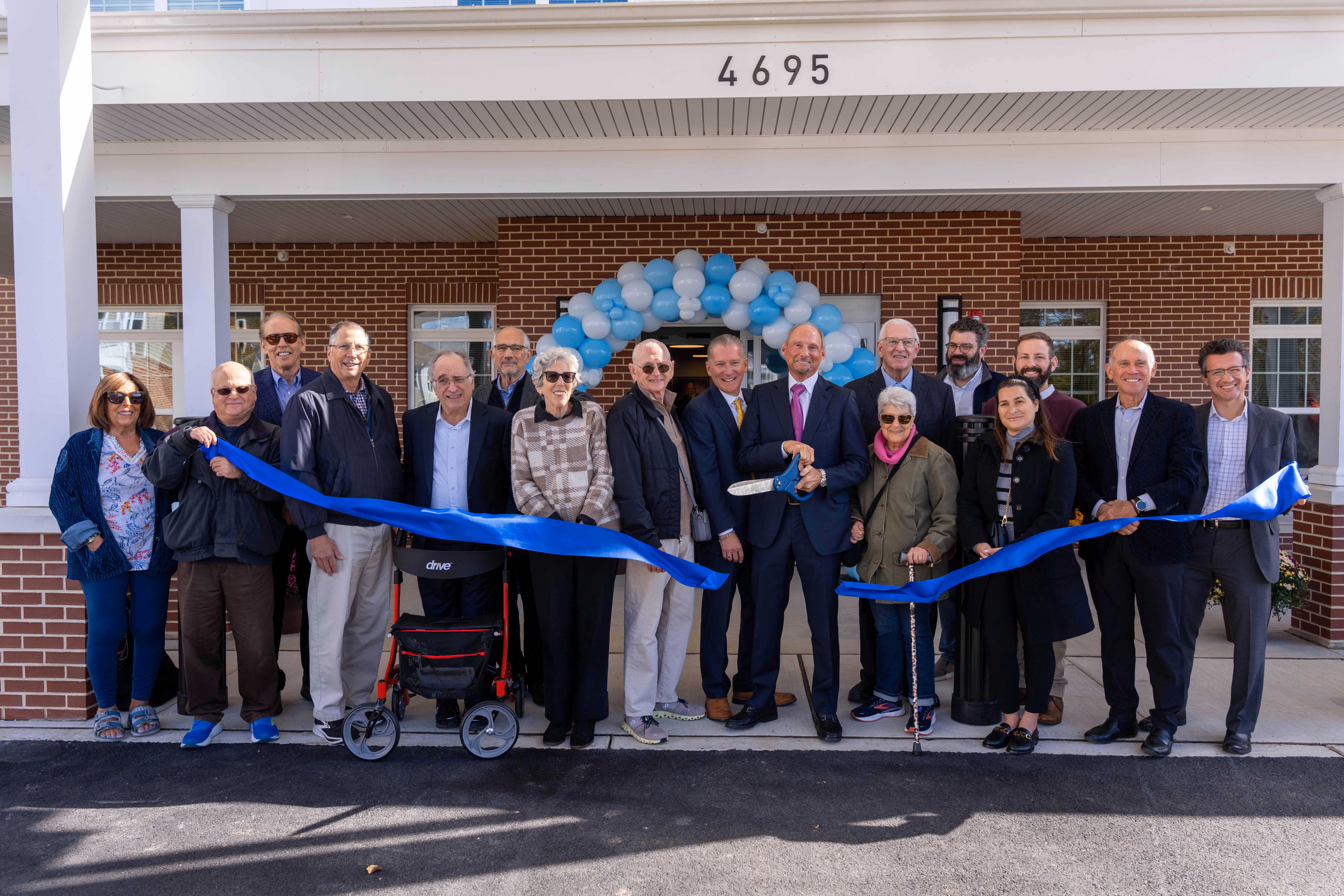 Group of people gathered outside a building entrance for a ribbon-cutting ceremony under a blue and white balloon arch.