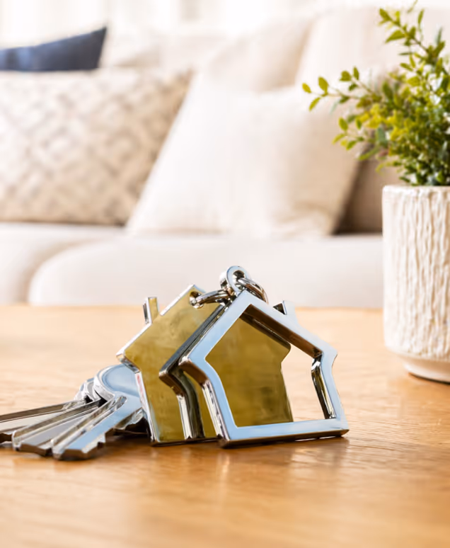 Keys with a house-shaped keychain on a wooden table with blurred couch and plant in the background.
