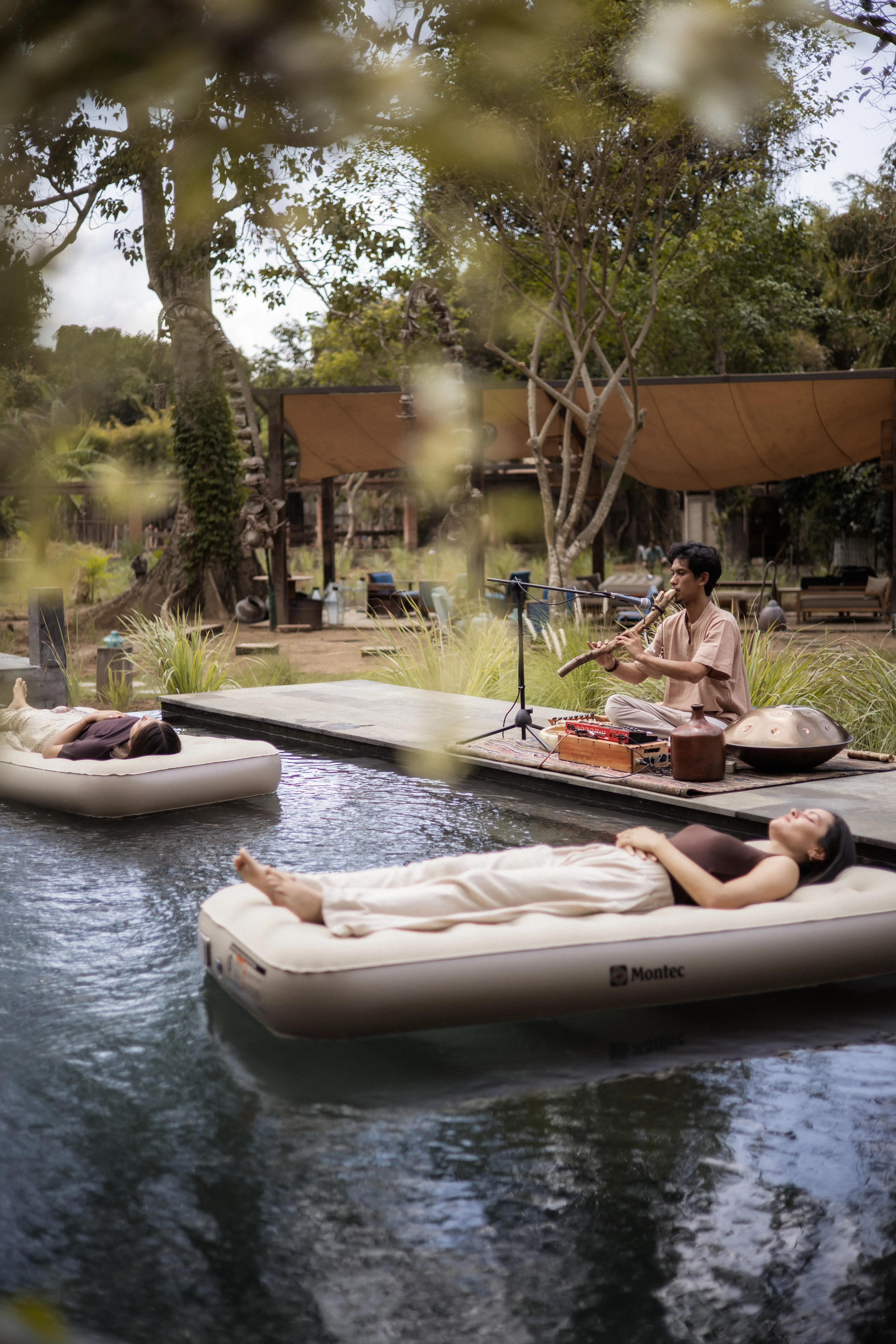 Participants floating on air mattresses in a pool while a musician plays a bamboo flute during an outdoor floating sound healing session.