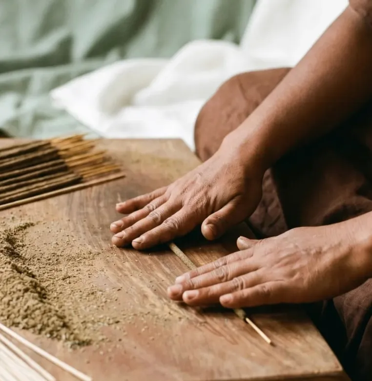 Hands rolling natural herbal powder onto a wooden stick over a timber table to craft handmade incense.