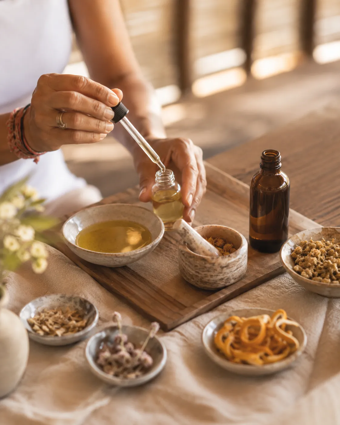 Close-up of hands using a dropper to blend essential oils into a glass bottle alongside bowls of dried herbs and botanicals.