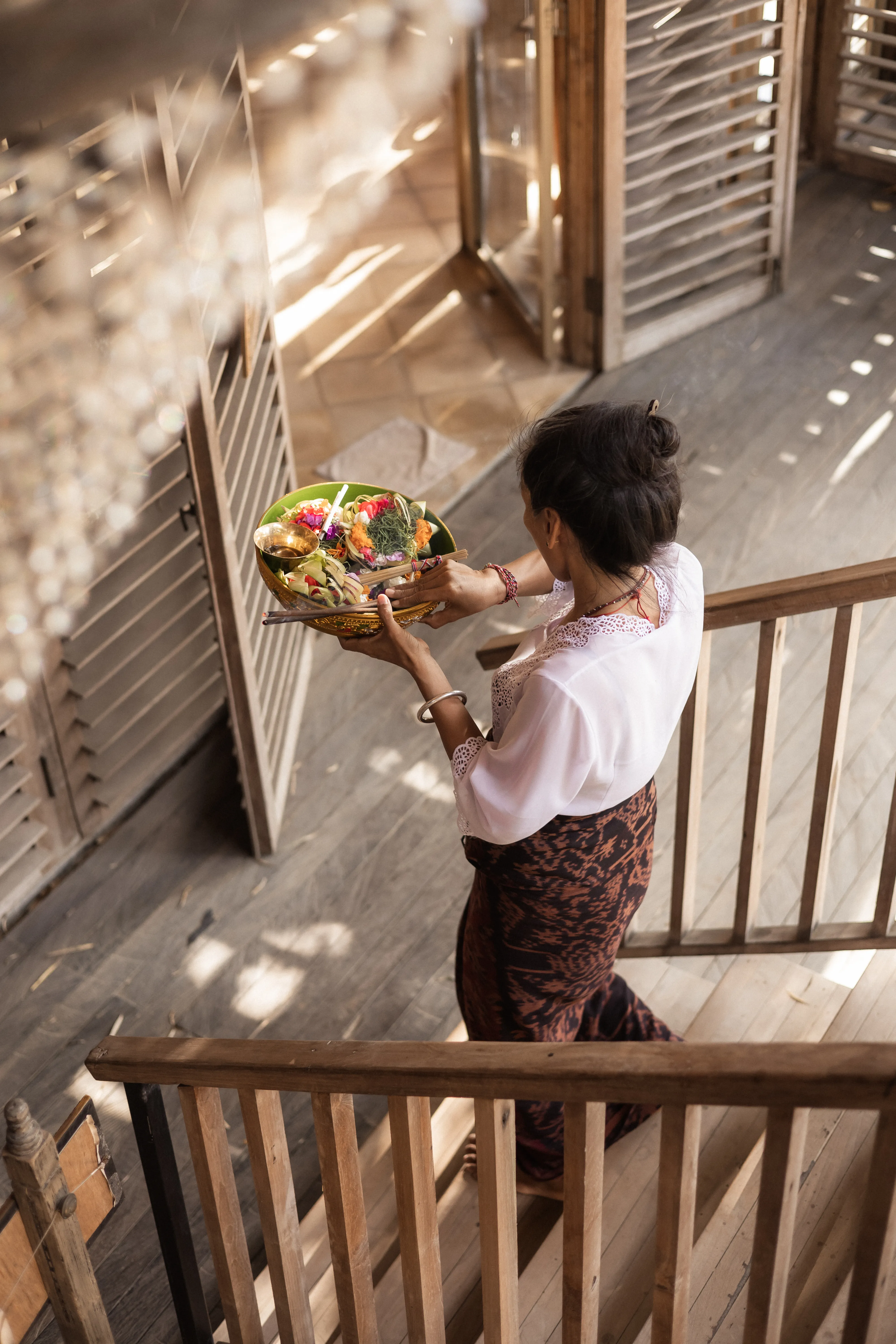 A Balinese woman in traditional attire walking up wooden steps while carrying a ceremonial tray of colorful flower offerings.