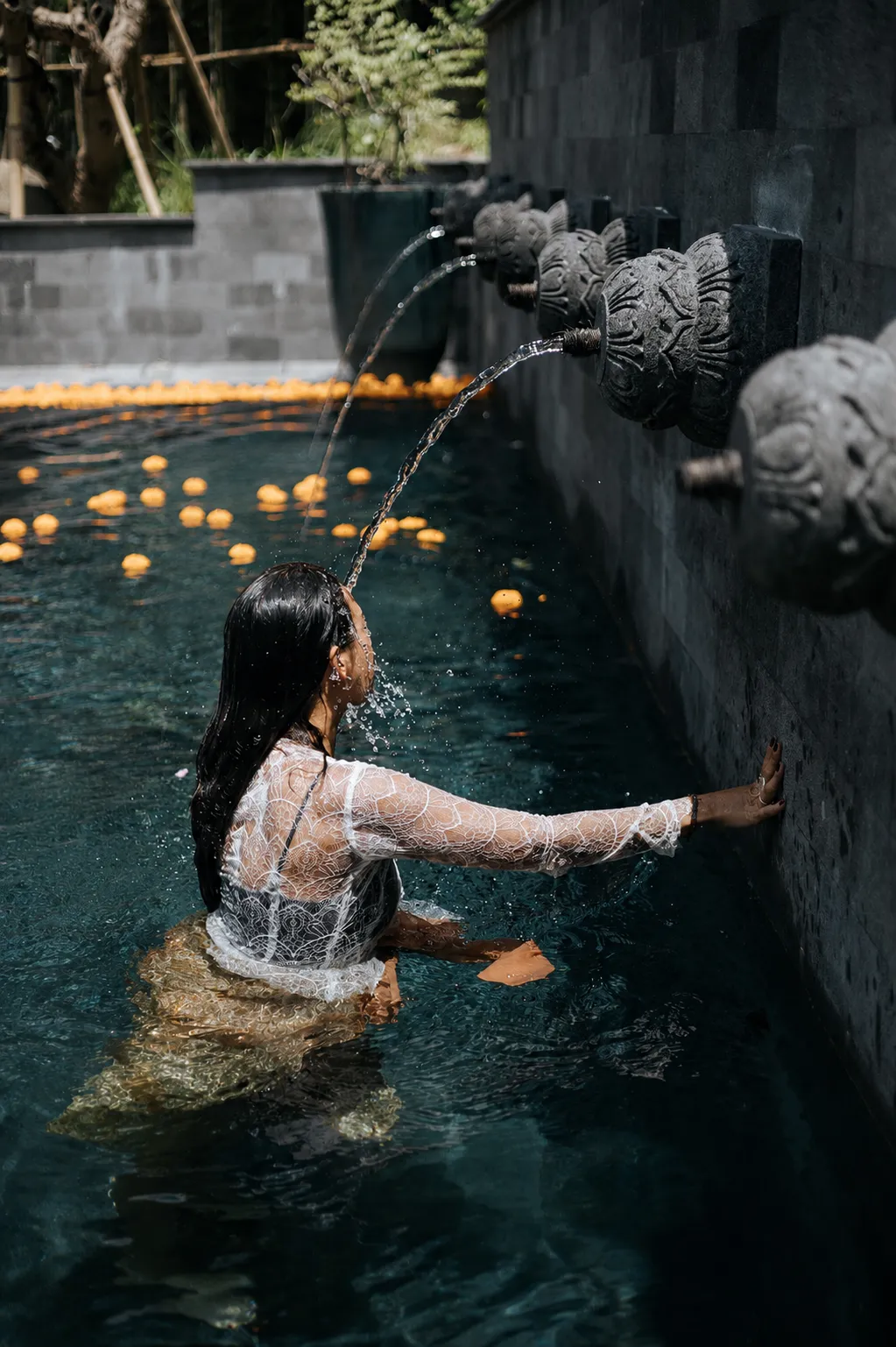 A woman receiving holy water purification under a carved stone fountain during a Balinese Melukat ceremony.