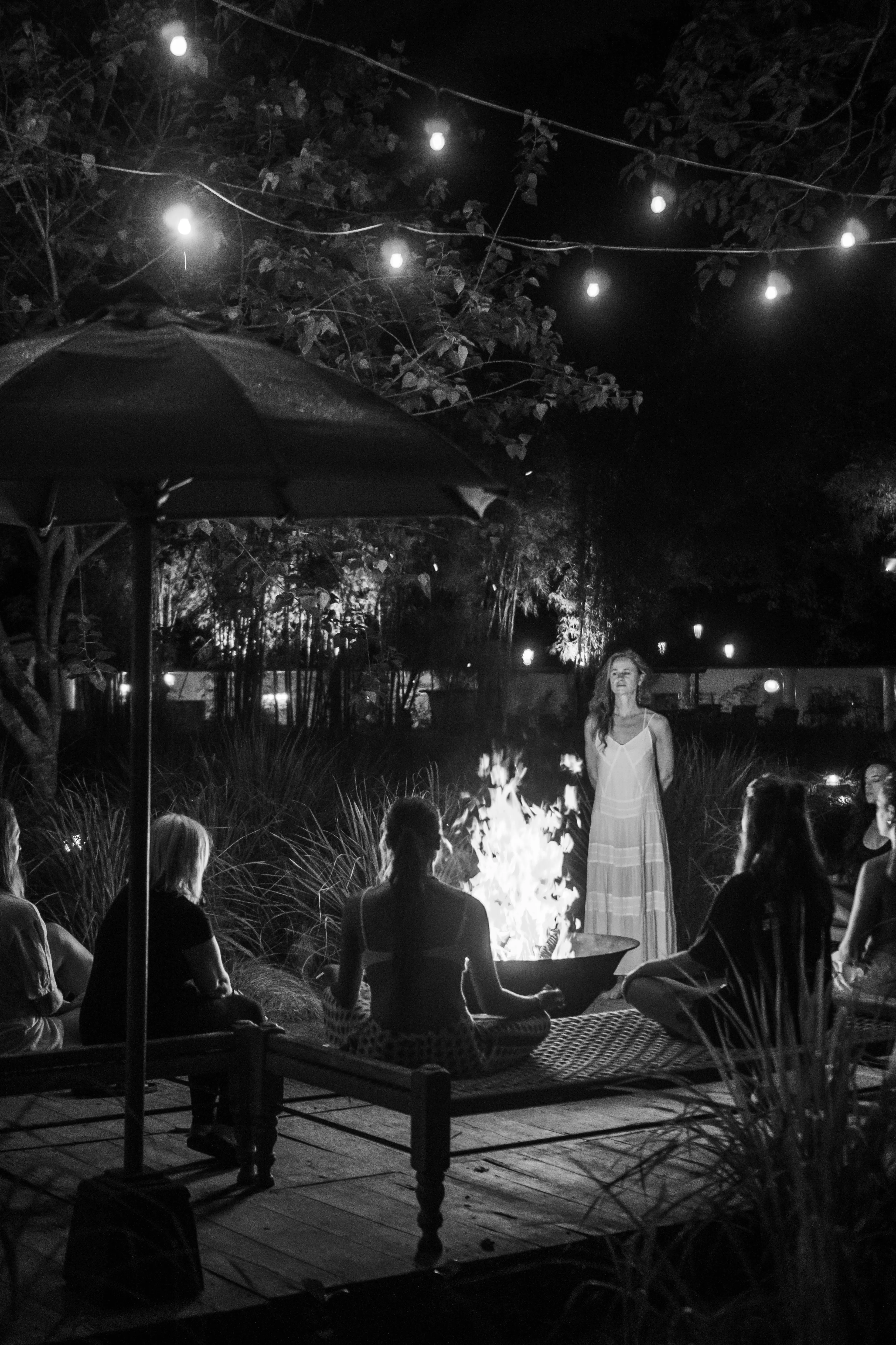 Black and white image of a facilitator and retreat participants gathered around a crackling outdoor campfire at night during a cacao ceremony.