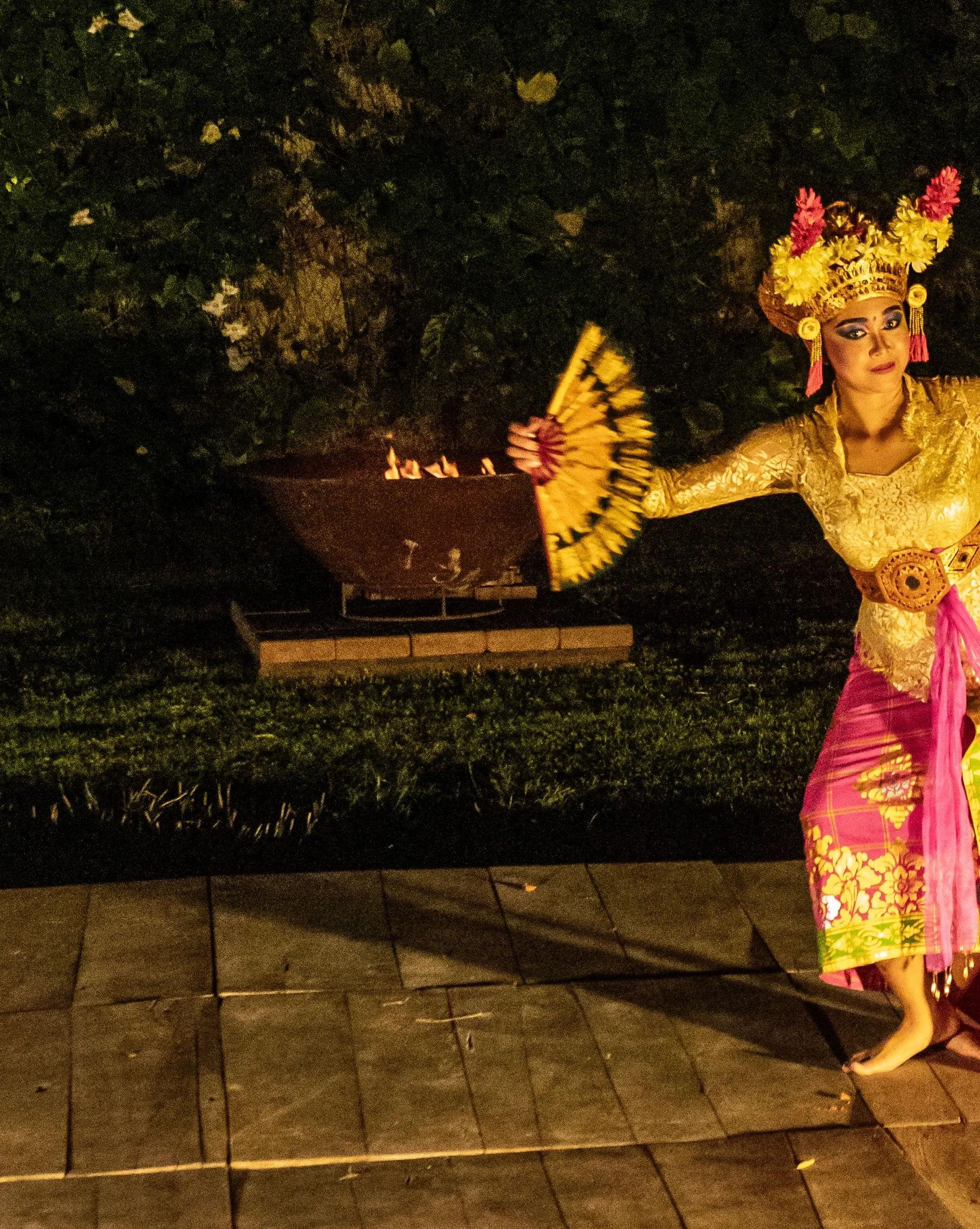 A Balinese dancer in vibrant gold costume and flower headdress holding a fan during an evening cultural performance near an open flame.