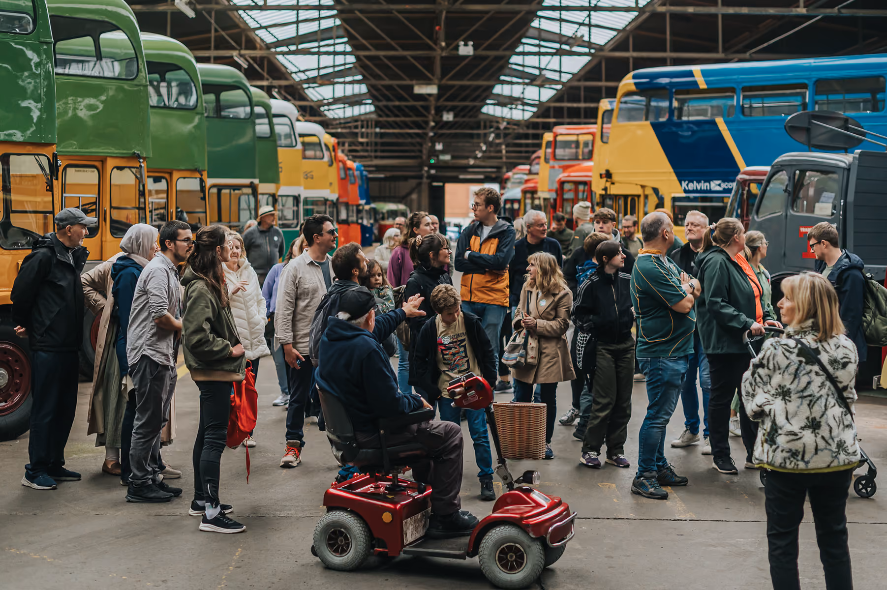 A large group listens to a tour guide in a vintage bus depot surrounded by colourful heritage double-decker buses