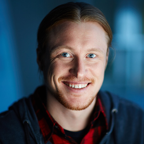Smiling man with blue eyes, red beard, and long hair tied back wearing a dark hoodie and red plaid shirt.