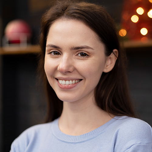 Smiling young woman with long brown hair wearing a light blue sweater in a softly lit room.
