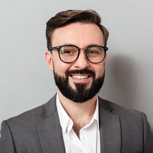 Smiling man with beard and glasses wearing a gray suit jacket and white shirt against a gray background.