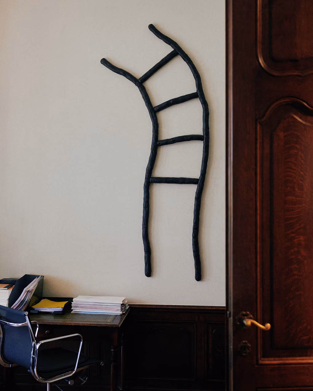 Moderne zwarte abstracte laddervormige wanddecoratie aan een witte muur in een kantoorruimte met een donker houten bureau, stapels papieren en een zwarte bureaustoel naast een deels geopend houten deur. Kunst te zien in het kantoor van Scriptas Notarissen.
