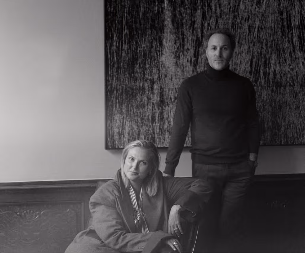 Zwart-witfoto van Notaris Melissa Saelens die in een stoel zit en Notaris Matthias Moortgat die naast haar staat tegen een muur met een groot kunstwerk.