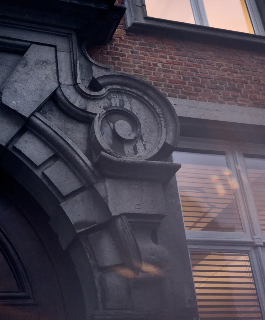 Close-up van een stenen architraaf met een gedetailleerd krulmotief boven een deur, met daarachter een bakstenen muur en ramen met jaloezieën.
