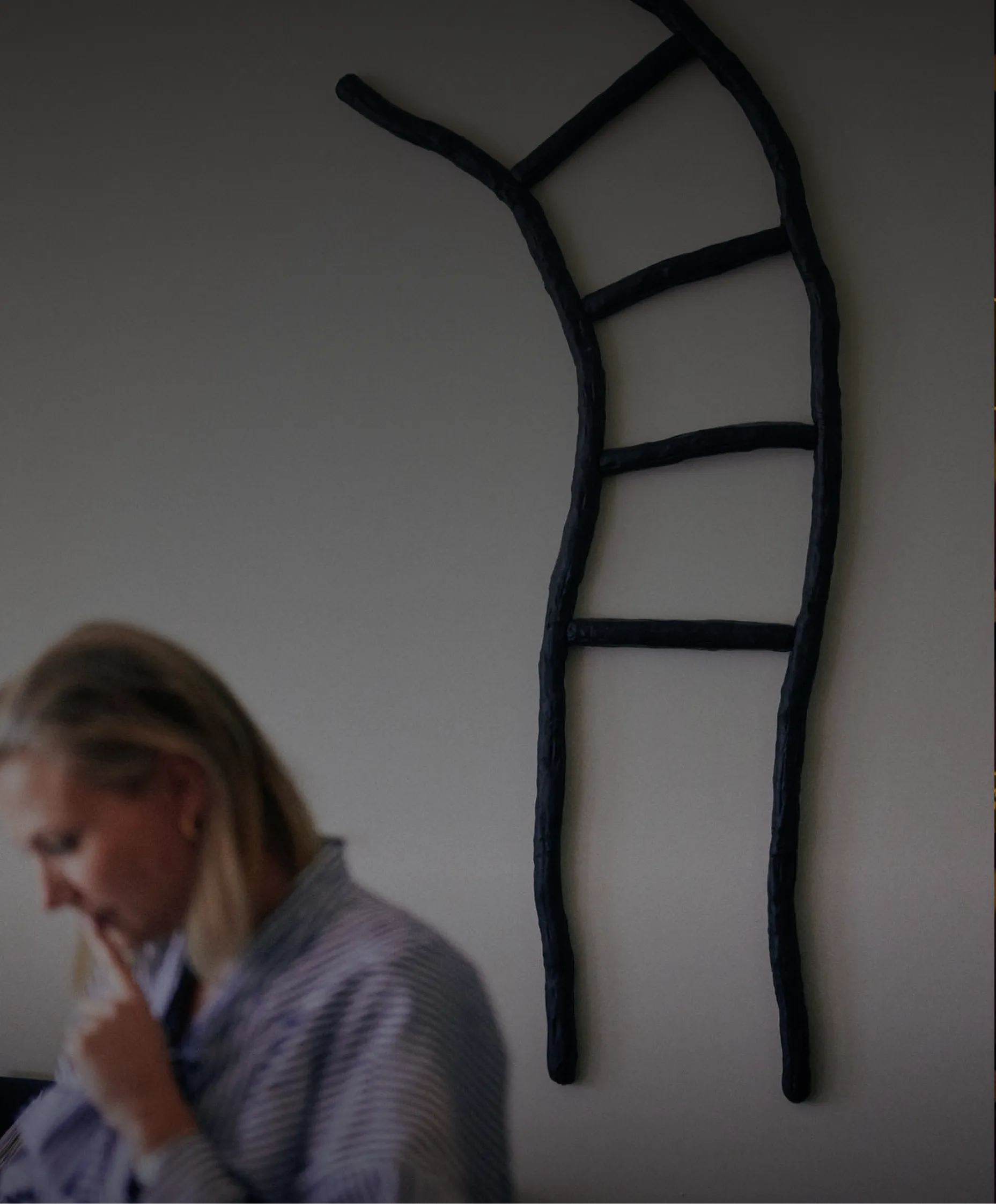 Vrouw, notaris Melissa Saelens, met blond haar en een gestreept overhemd zit nadenkend met haar vinger bij haar mond, en een abstracte zwarte ladder met vier sporten aan de muur achter haar.