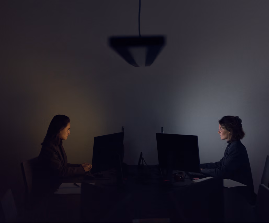 Twee vrouwen zitten aan tegenovergestelde kanten van een tafel, elk werkend op een computer in een donkere kamer met een enkele hanglamp boven hen.