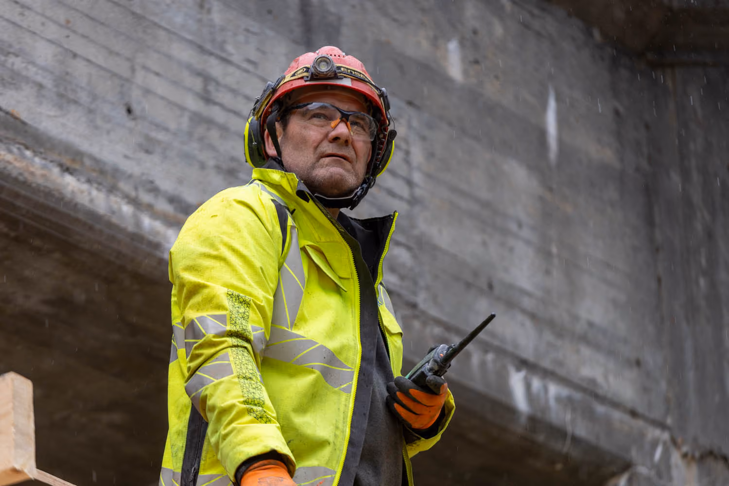 En bygningsarbeider i neongul sikkerhetsjakke, hjelm og hansker holder en walkie-talkie foran en betongkonstruksjon.