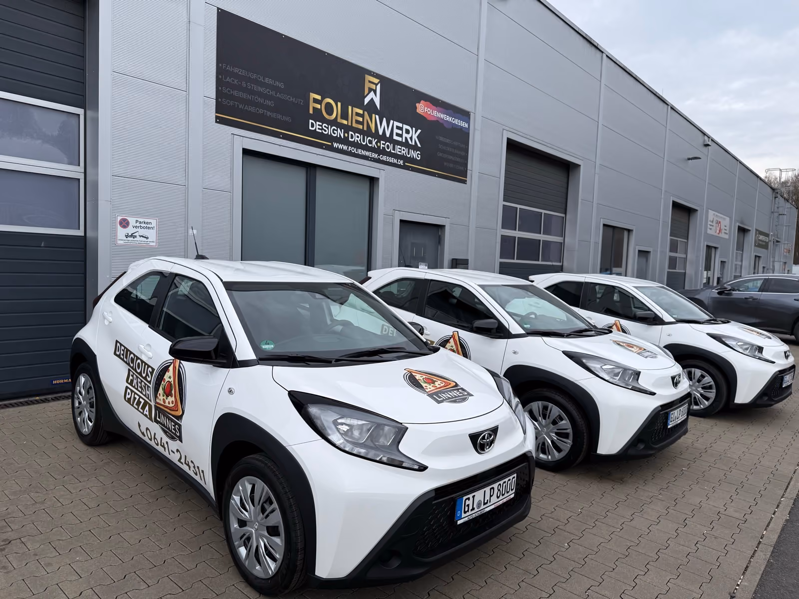 Drei weiße Toyota Aygo X mit Werbebeschriftung eines Pizza-Lieferdiensts vor dem Folienwerk Gießen