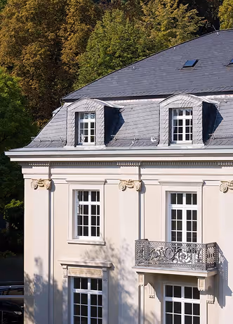 Part of a cream-colored building with classic windows, balcony, and a slate roof under trees.