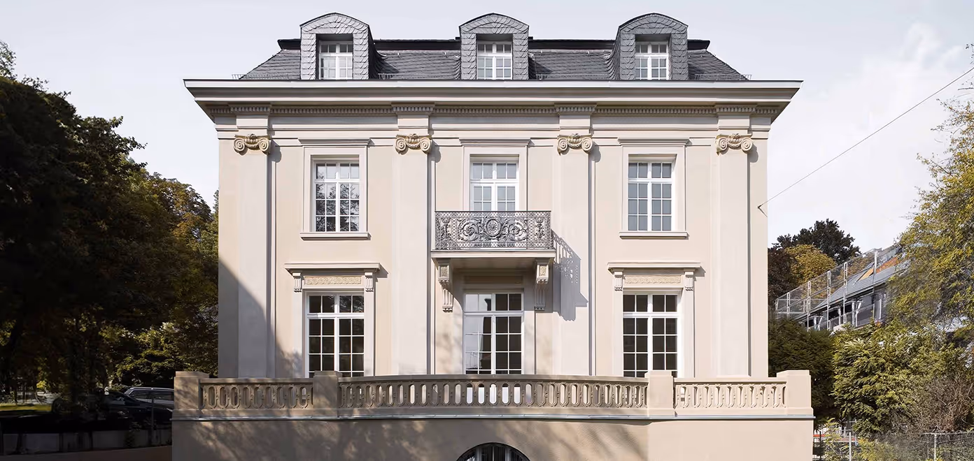 Front view of a classic beige mansion with tall windows, decorative balcony, and dormer windows on roof.