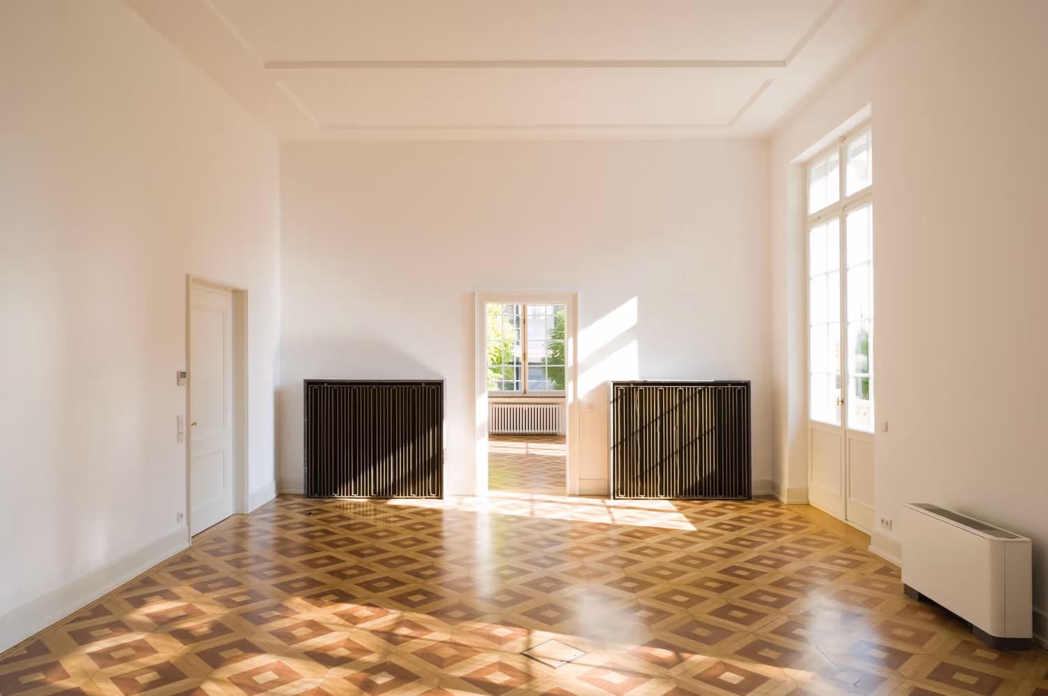 Empty room with patterned wooden floor, two black radiators, white walls, and large windows.