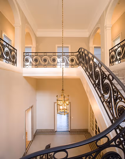 Curved staircase with intricate black iron railing and hanging chandelier in beige interior.